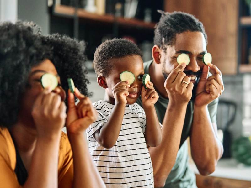 Mutter und Vater albern mit ihrem Sohn in der Küche herum. Alle halten sich Zucchini-Scheiben vor die Augen.