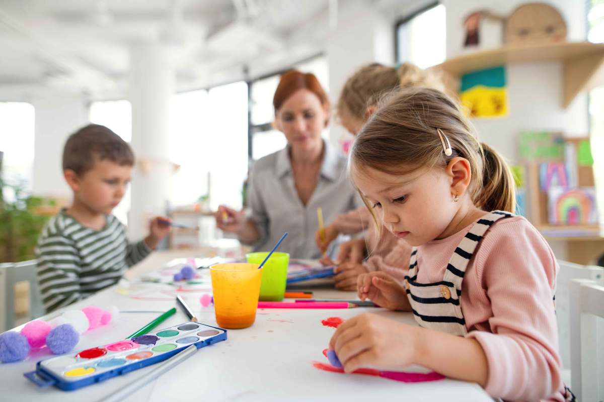 Drei Vorschulkinder sitzen in ihrem Gruppenraum mit einer Erzieherin am Tisch und malen mit Wasserfarben.