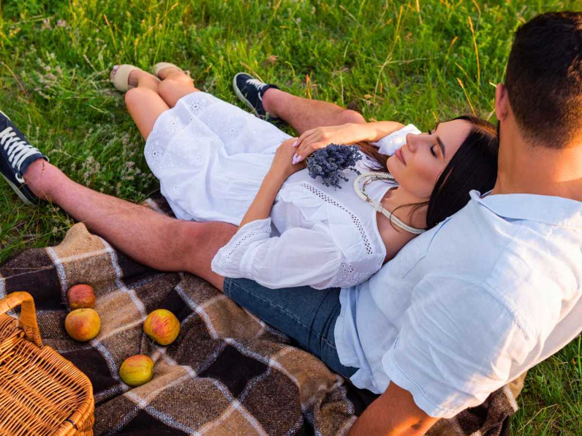 Frau mit geschlossenen Augen und Lavendelbl&uuml;ten entspannt neben ihrem Freund und &Auml;pfeln auf einer karierten Decke.