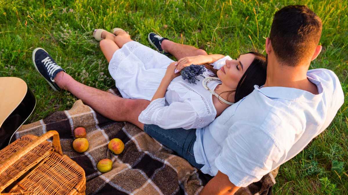 Frau mit geschlossenen Augen und Lavendelblüten entspannt neben ihrem Freund und Äpfeln auf einer karierten Decke.