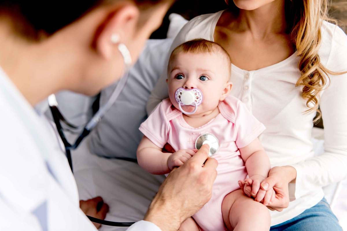 Baby sitzt auf dem Schoß der Mutter und schaut den Arzt mit großen Augen an, der es mit einem Stethoskop abhören möchte,