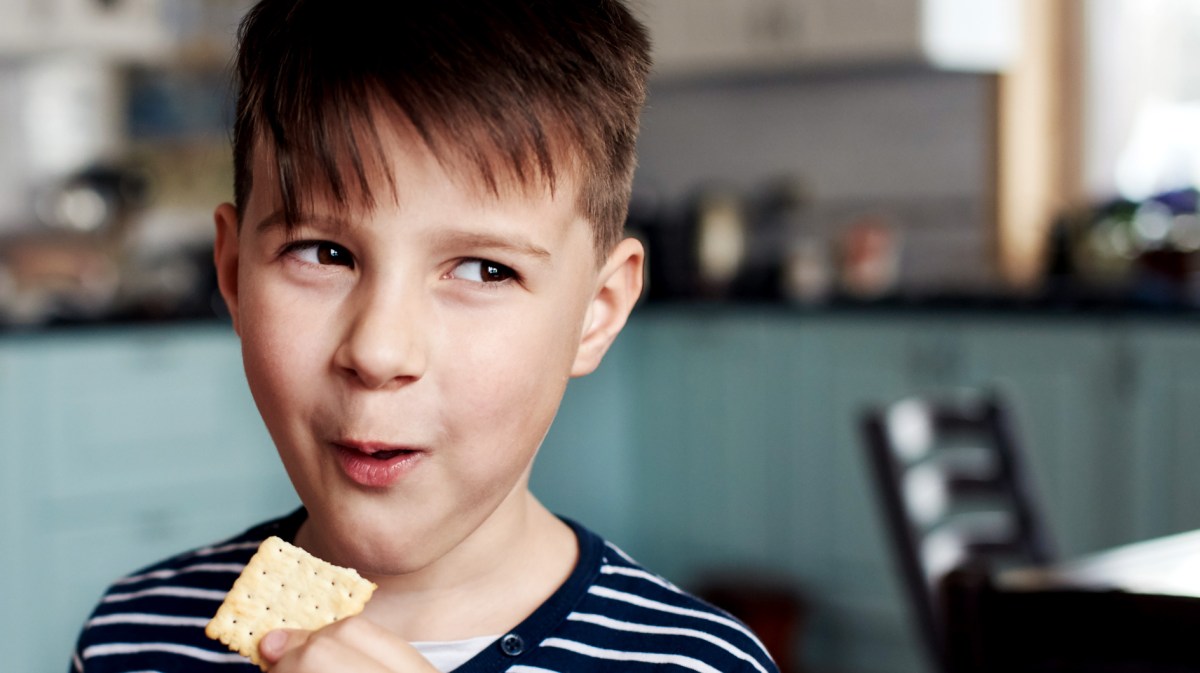 Junge, ca. 10 Jahre alt, schaut verschmitzt zur Seite und isst dabei einen Cracker.