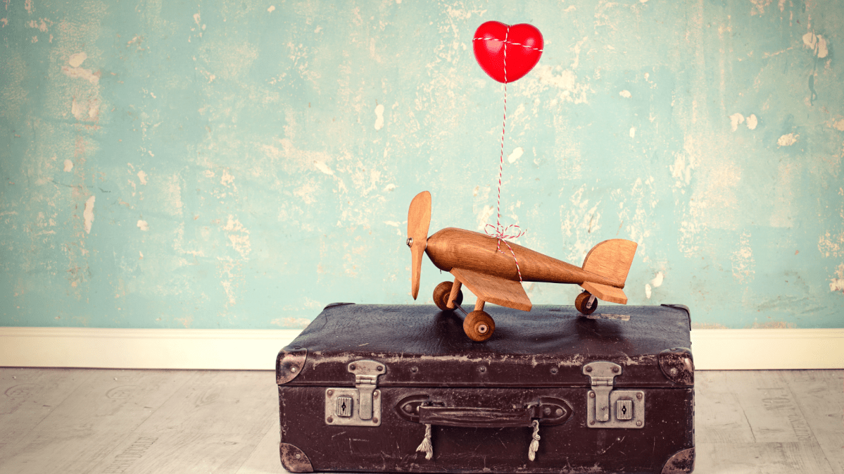 Auf einem alten brauen Koffer stejt ein kleines gelbes Flugzeug mit einem roten Herzluftballon angehängt.