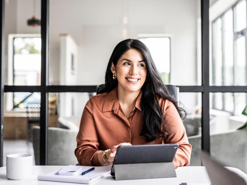 Frau mit dunklen Haaren sitzt am Schreibtisch im Büro an einem Tablet.