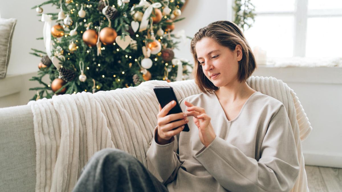 Frau sitzt mit einem Handy auf der Couch vor dem Weihnachtsbaum.