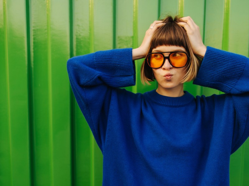 Frua mit Brille und blauem Pullover, die sich in die Haare fasst und vor einer grünen Wand steht