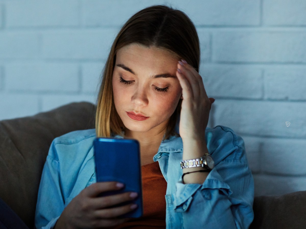 Frau auf einer Couch, die sich an die Schl&auml;fe packt und nachdenklich in ihr Handy schaut