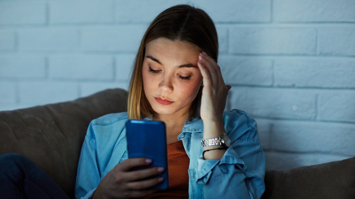 Frau auf einer Couch, die sich an die Schläfe packt und nachdenklich in ihr Handy schaut