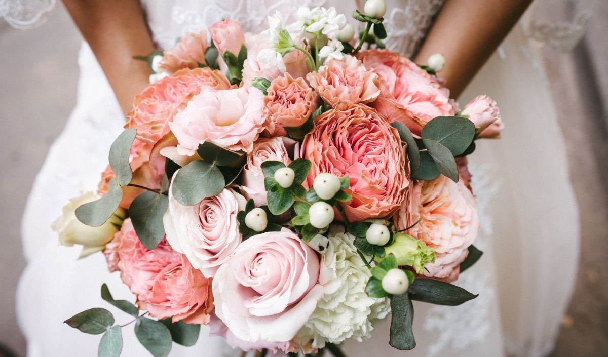 Braut hält einen Brautstrauß mit Rosen und Pfingstrosen vor sich.