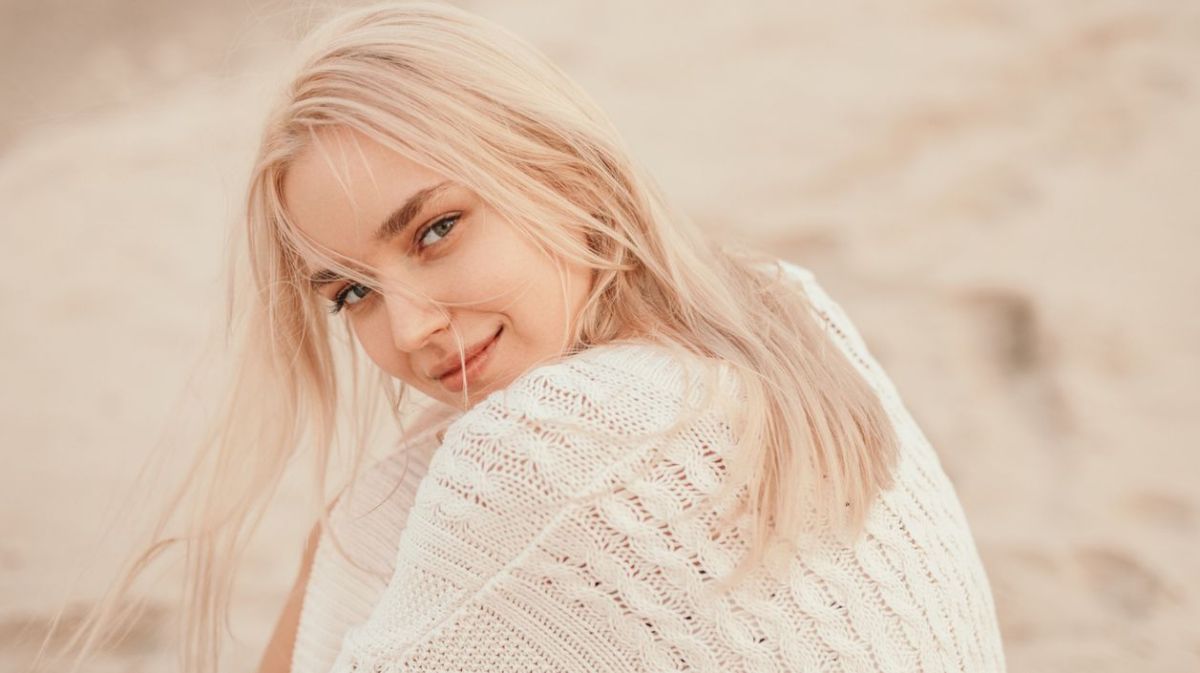 Frau mit blonden Haaren sitzt am Strand und schaut in die Kamera.