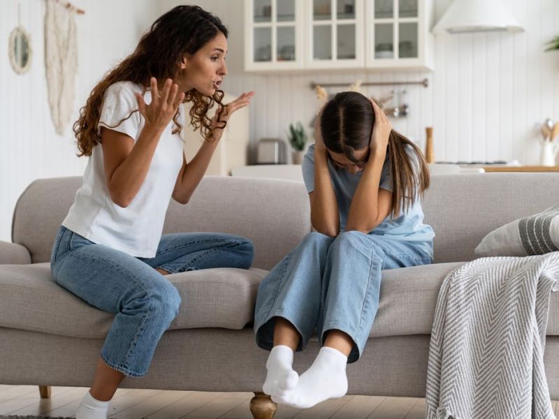 Mutter und Tochter sitzen auf dem Sofa im Wohnzimmer. Die Mutter schimpft mit ihrer Tochter, während die sich die Ohren zuhält.