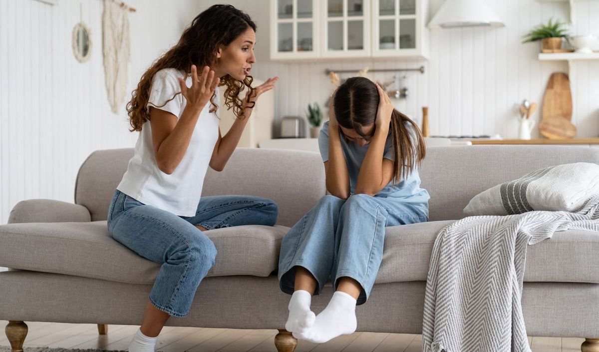 Mutter und Tochter sitzen auf dem Sofa im Wohnzimmer. Die Mutter schimpft mit ihrer Tochter, während die sich die Ohren zuhält.