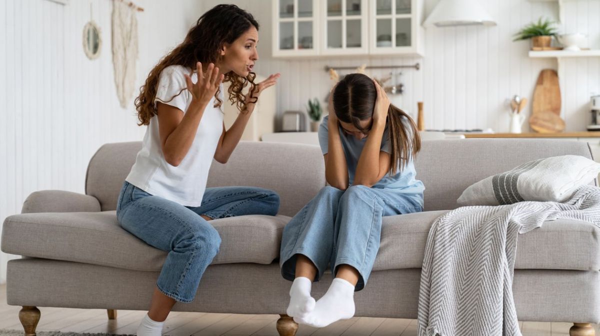 Mutter und Tochter sitzen auf dem Sofa im Wohnzimmer. Die Mutter schimpft mit ihrer Tochter, während die sich die Ohren zuhält.