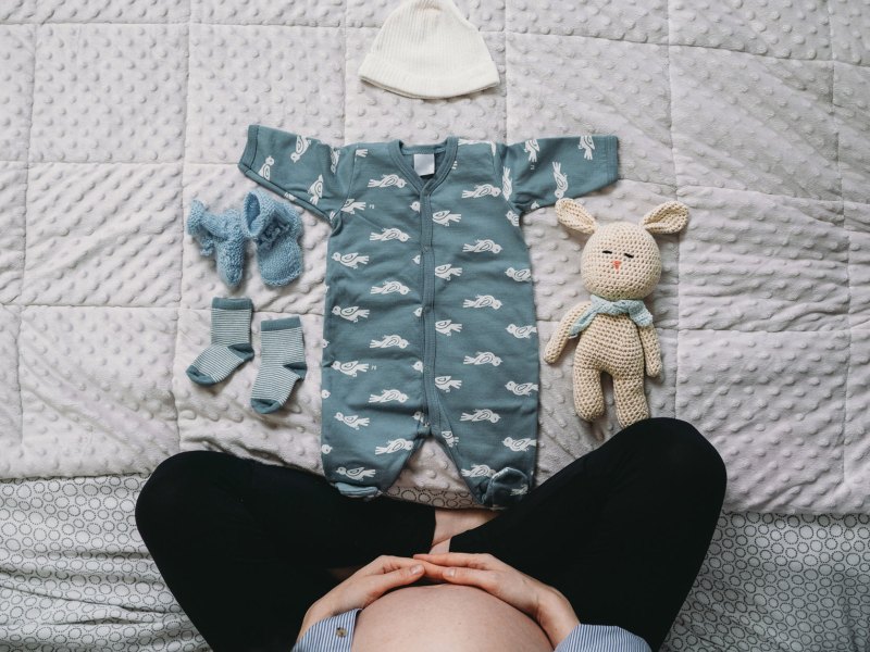 Schwangere sitzt auf dem Bett, vor sich Babykleidung