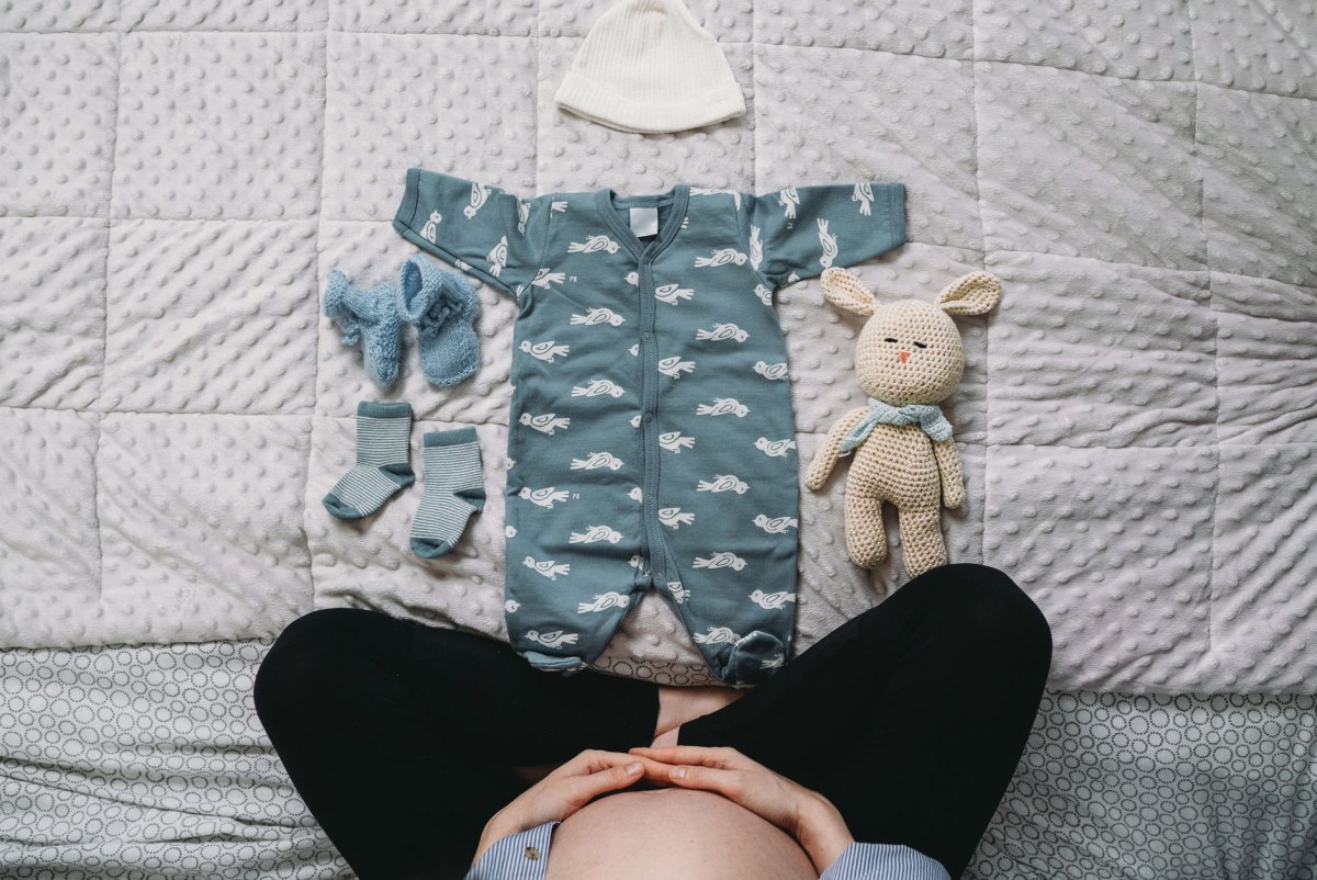 Schwangere sitzt auf dem Bett, vor sich Babykleidung