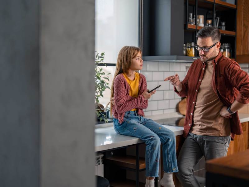Vater schimpft mit seiner Tochter, die trotzig und mit einem Handy in der Hand auf der Küchenzeile sitzt.