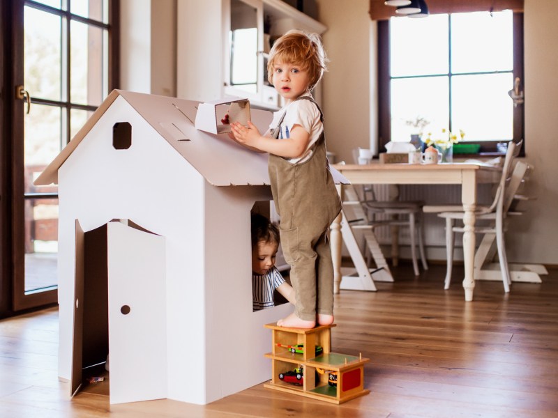 Zwei kleine Kinder spielen drinnen mit einem Spielhaus. Ein Kind schaut von innen aus dem Spielhaus heraus, eines steht auf einer kleinen Treppe daneben.