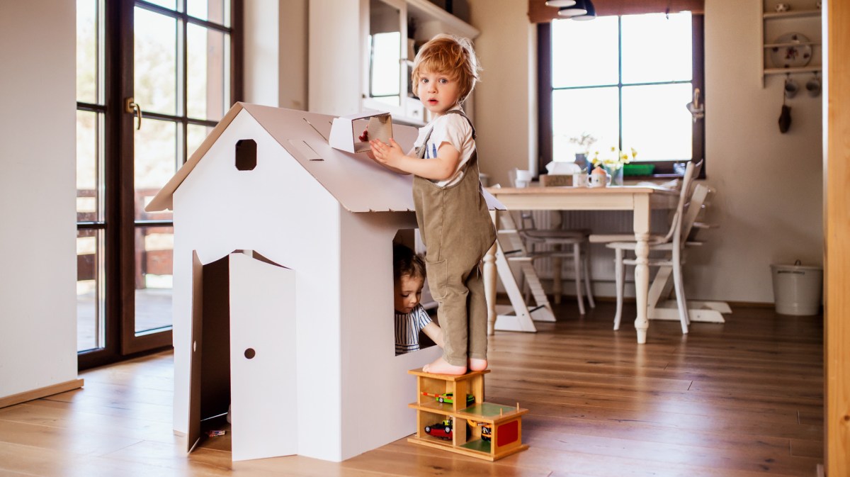 Zwei kleine Kinder spielen drinnen mit einem Spielhaus. Ein Kind schaut von innen aus dem Spielhaus heraus, eines steht auf einer kleinen Treppe daneben.