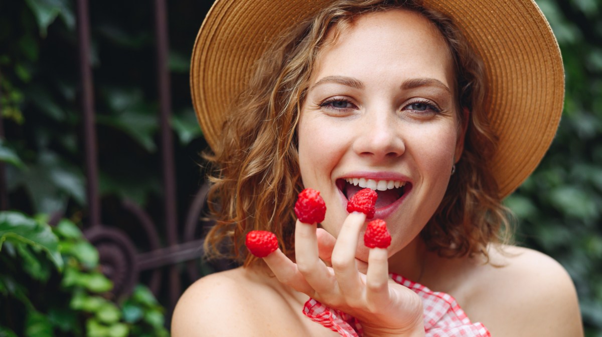 Frau mit Hut, hat sich Himbeeren auf die Fingerspitzen gesteckt.