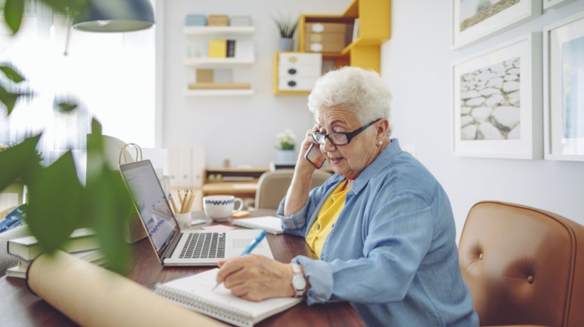 Rentnerinnen sitzt wegen einem Minijob am Laptop und telefoniert.