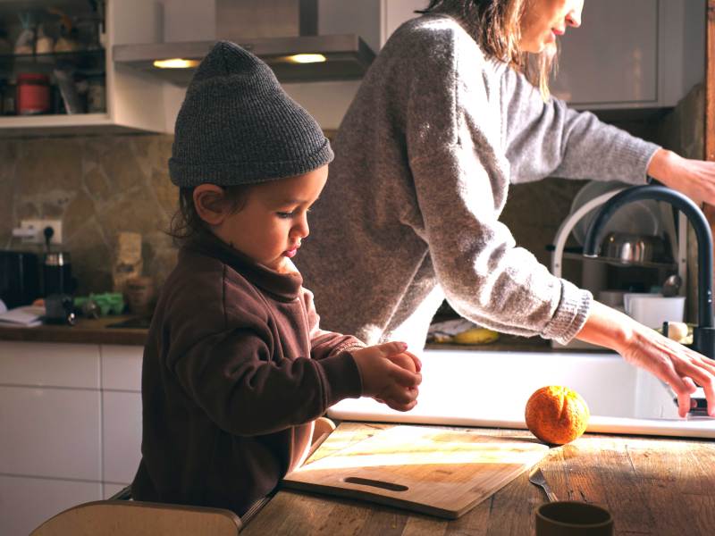 Kleiner Junge steht konzentriert an der Küchenzeile und schält sich allein eine Orange. Die Mutter wäscht daneben ab.