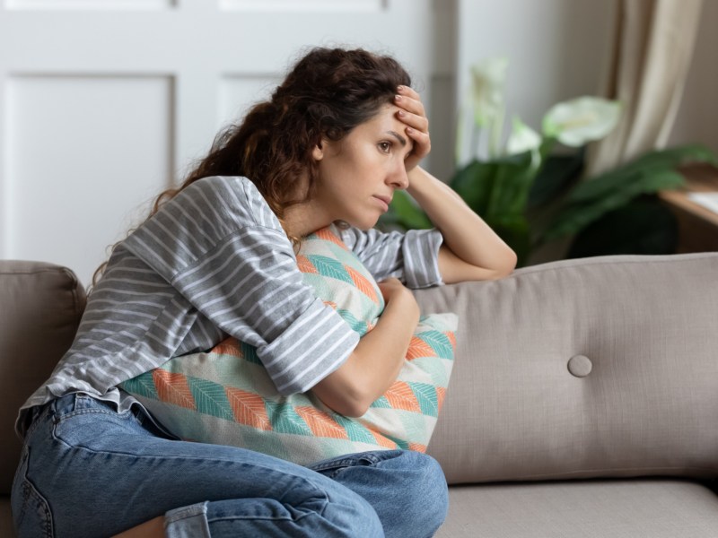 Frau auf dem Sofa, die mit einem Kissen auf dem Beinen liegt und sich nachdenklich an den Kopf packt