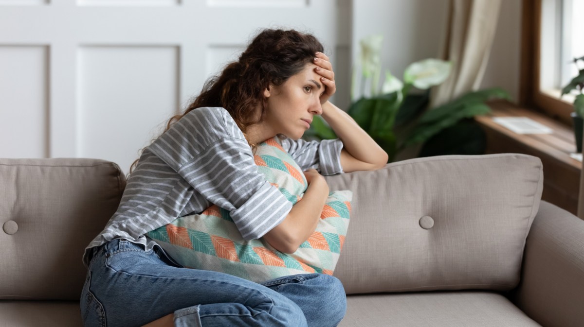 Frau auf dem Sofa, die mit einem Kissen auf dem Beinen liegt und sich nachdenklich an den Kopf packt