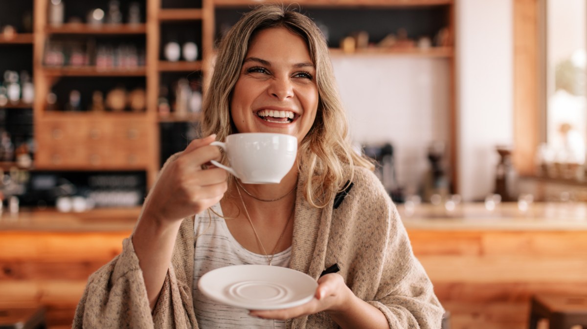 Frau mit blonden Haaren lacht und hält Kaffeetasse in Hand.