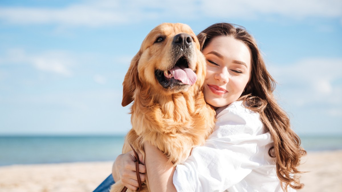 Frau kuschelt Hund am Strand.