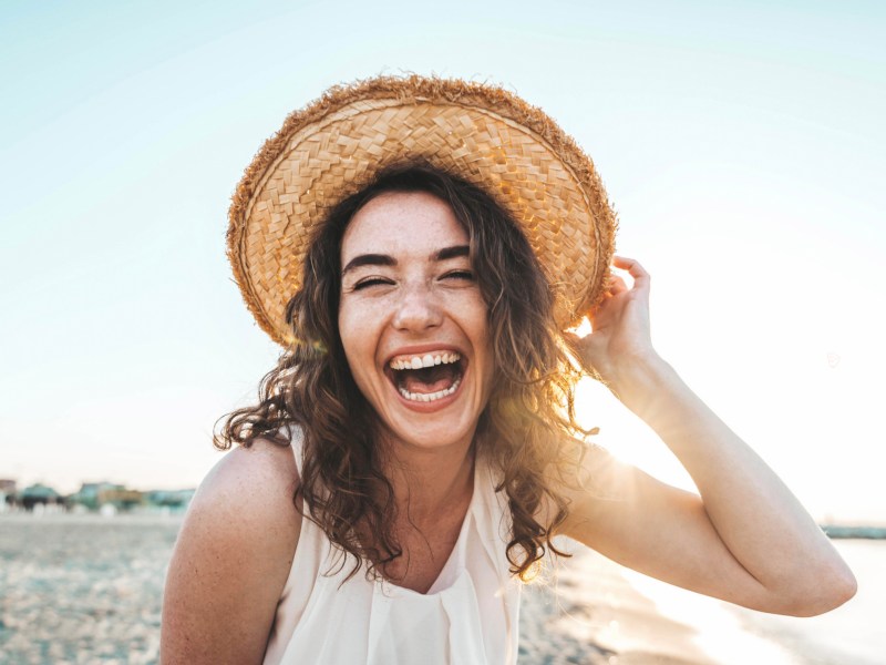 Frau am Strand mit Hut auf dem Kopf, die lächelnd in die Kamera blickt.