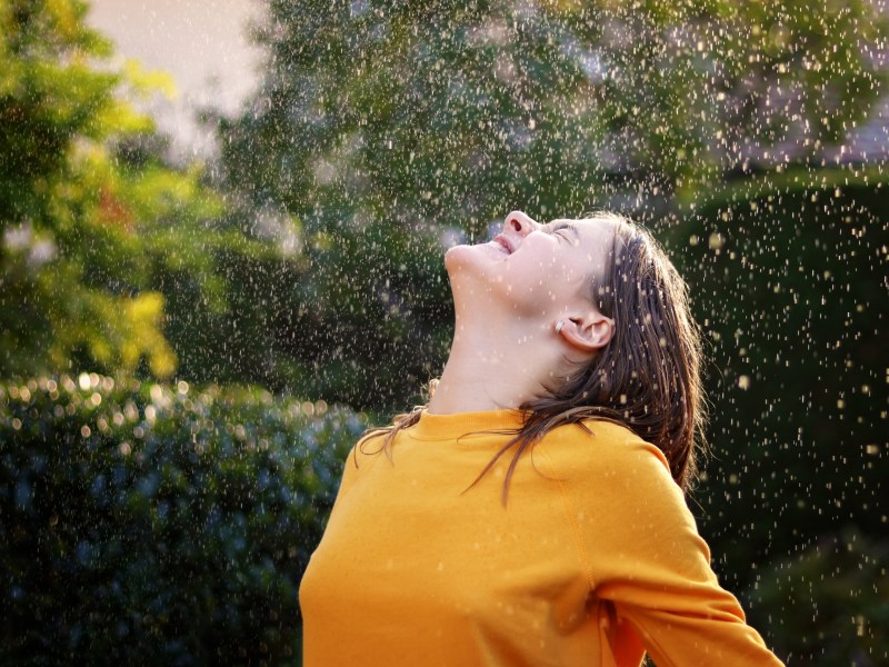 Frau steht im Sommerregen und streckt lächelnd das Gesicht zum Himmel.