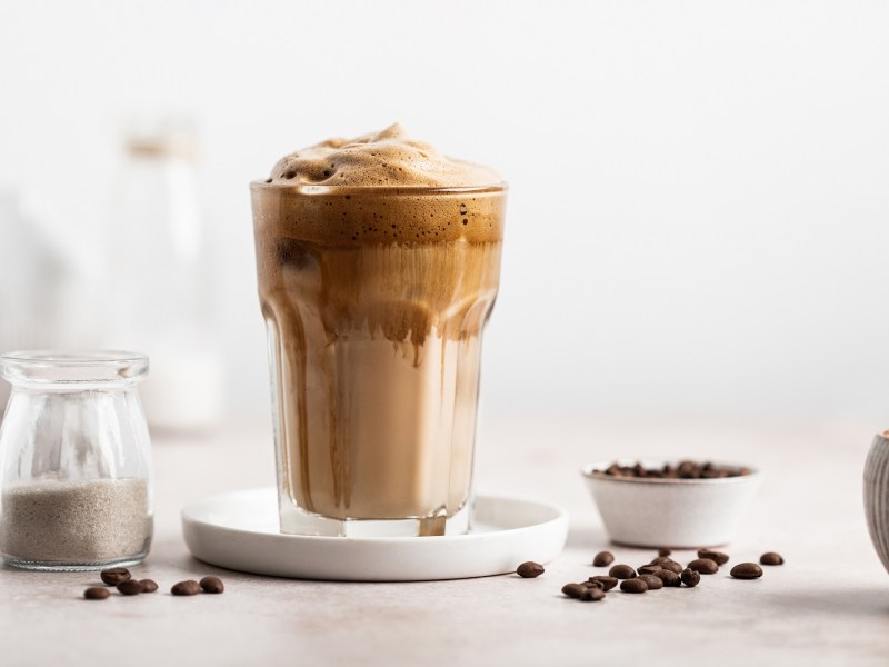 Ein Glas griechischer Frappé mit Kaffeeschaum, daneben Kaffeebohnen und ein Kännchen mit Milch.