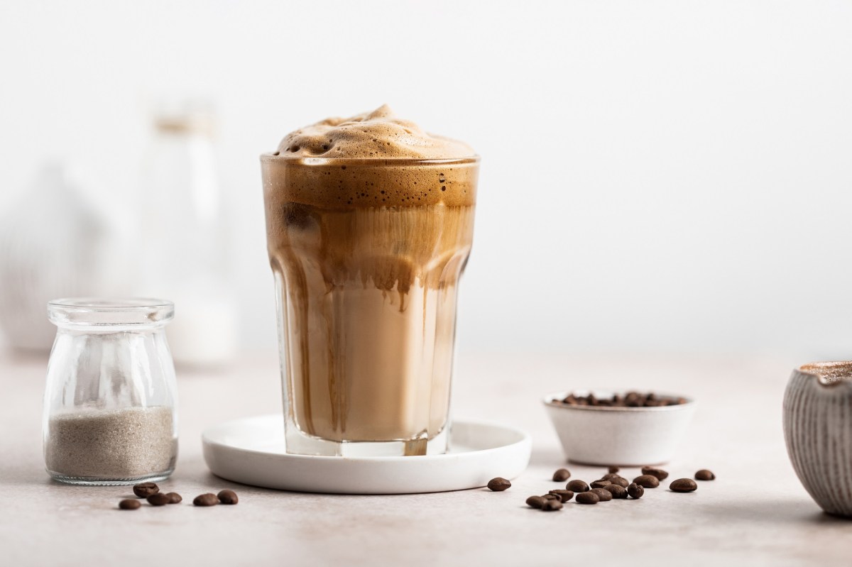 Ein Glas griechischer Frappé mit Kaffeeschaum, daneben Kaffeebohnen und ein Kännchen mit Milch.