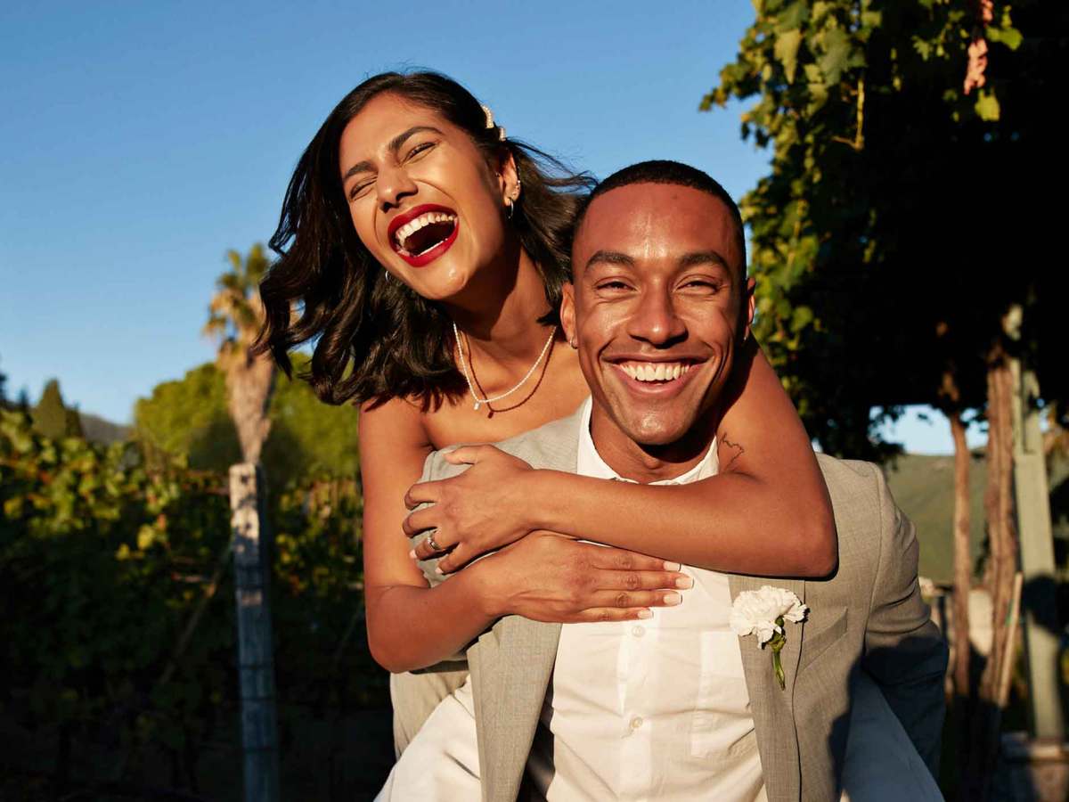 Braut h&auml;ngt auf dem R&uuml;cken ihres Ehemannes. Beide lachen ausgelassen.