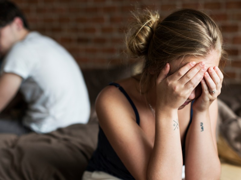 Frau sitzt mit Händen vor dem Gesicht auf Bettkante mit verschwommenen Bild eines Mannes im Hintergrund