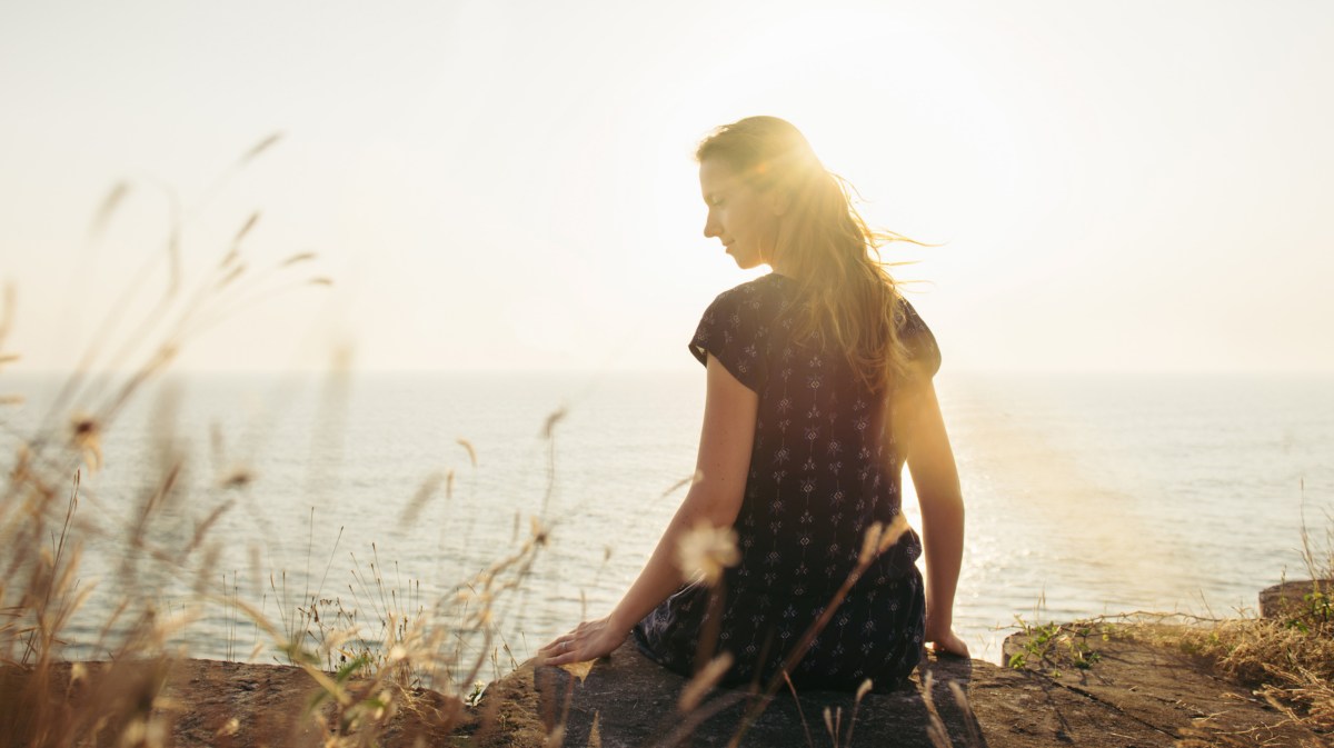 Frau an Klippe am Meer, die durch die Sonne nach hinten in die Kamera blickt