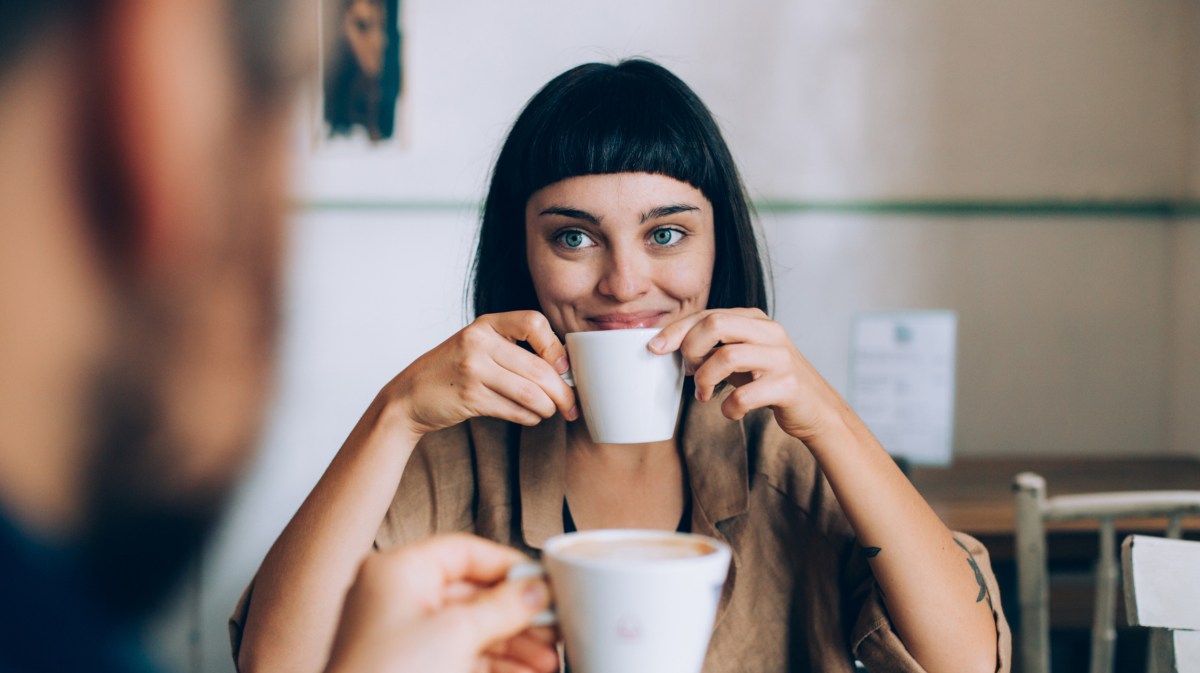 Frau im Café, die lacht und an Cappuccino nippt, mit unscharfem Mann im Vordergrund, der anstoßen möchte