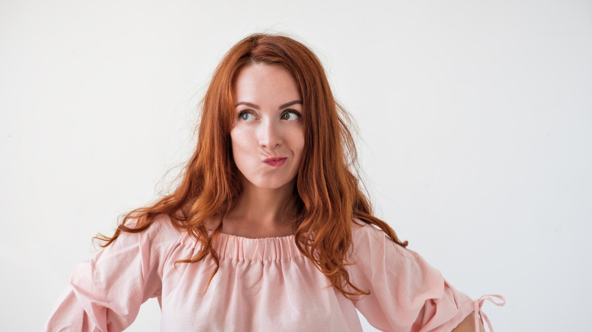 Frau mit roten Haaren guckt mit Schnute nach links vor weißer Wand