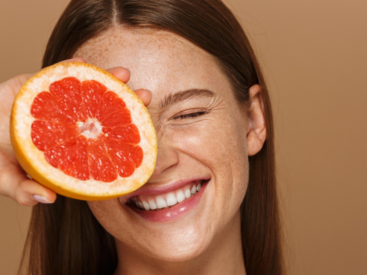 Frau vor beigem Hintergrund mit Sommersprossen, die sich eine Grapefruit vors Gesicht hält und lacht