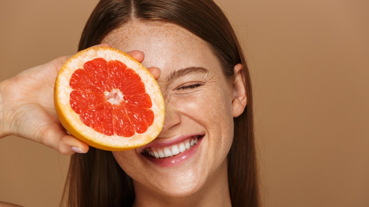 Frau vor beigem Hintergrund mit Sommersprossen, die sich eine Grapefruit vors Gesicht hält und lacht