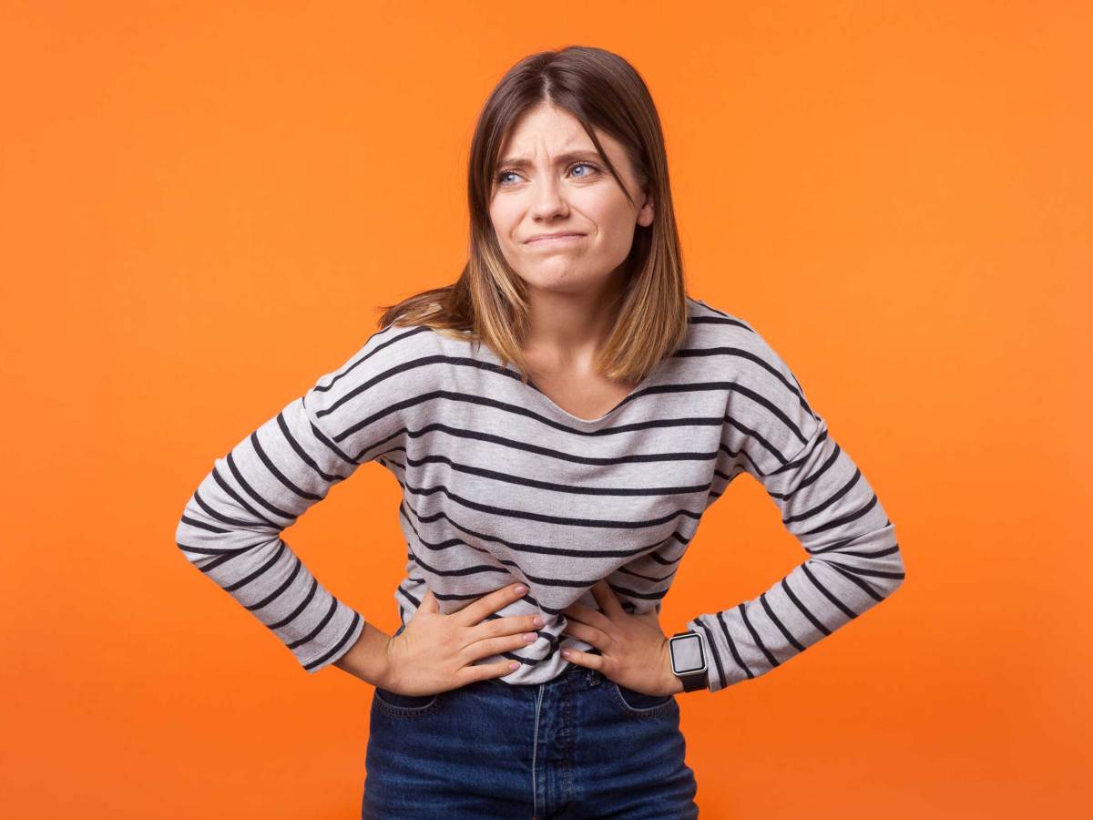 Porträt einer unglücklichen kranken Frau mit braunen Haaren in einem langärmeligen gestreiften Hemd, die ihren Bauch mit den Händen festhält, Magenkrämpfe oder Menstruationsschmerzen. Innenaufnahme im Studio, isoliert auf orangefarbenem Hintergrund