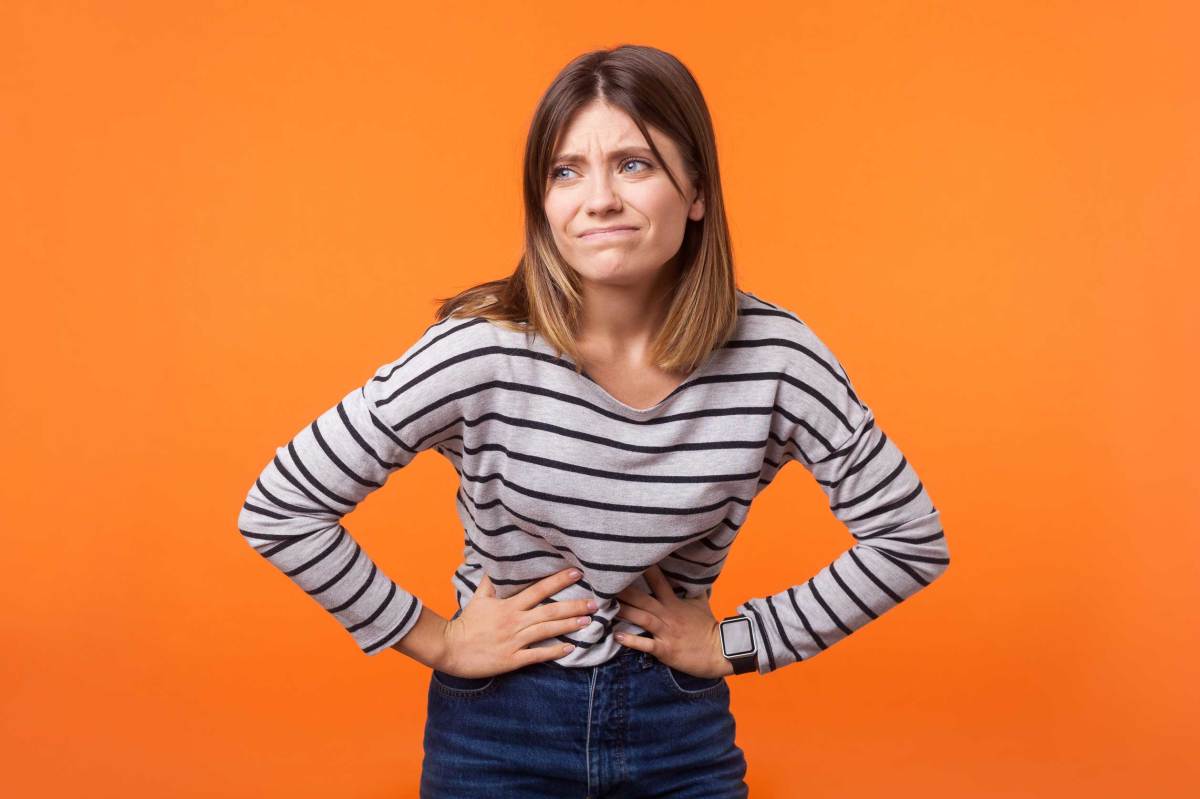 Porträt einer unglücklichen kranken Frau mit braunen Haaren in einem langärmeligen gestreiften Hemd, die ihren Bauch mit den Händen festhält, Magenkrämpfe oder Menstruationsschmerzen. Innenaufnahme im Studio, isoliert auf orangefarbenem Hintergrund