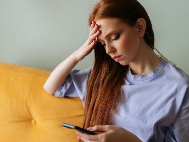 Rothaarige Frau auf gelber Couch, die nachdenklich auf Handy schaut