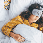 Millennial-Frau im Bett liegend mit Schlafmaske und Telefon in der Hand.