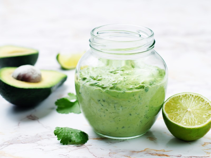 Tzatziki mit Avocado in einem Glas, daneben halbierte Avocados und Limetten.