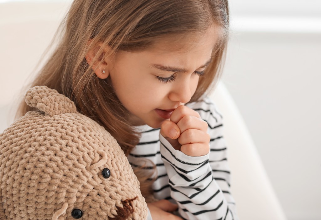 Kleines Mädchen mit Teddy im Arm hustet in ihre Faust.