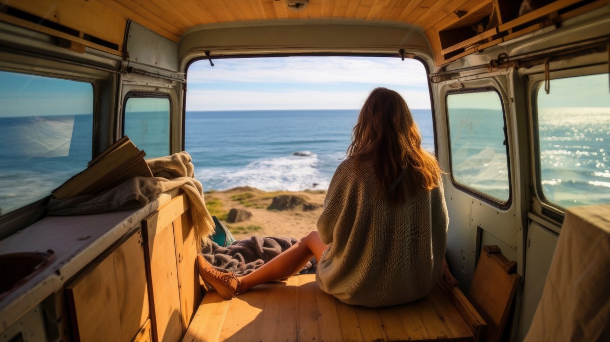 Frau in einem Van, die im Kofferraum in einem T-Shirt sitzt und auf das Meer und die Klippen schaut