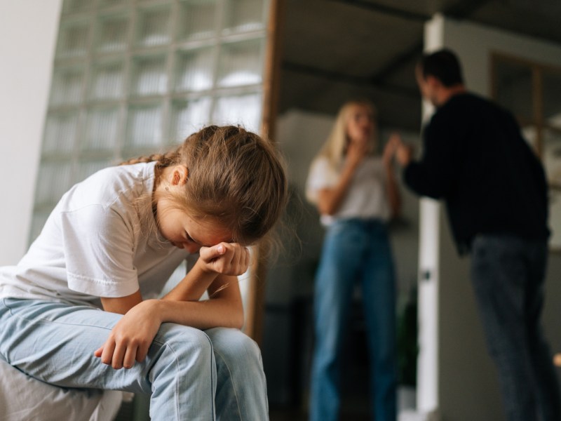 Zwei Erwachsene, die sich im Hintergrund streiten, während ein Kind im Vordergrund ihre Hände vors Gesicht nimmt