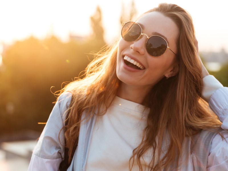 Frau mit Sonnenbrille geht sich durch die Haare und steht lachend in der Sonne.