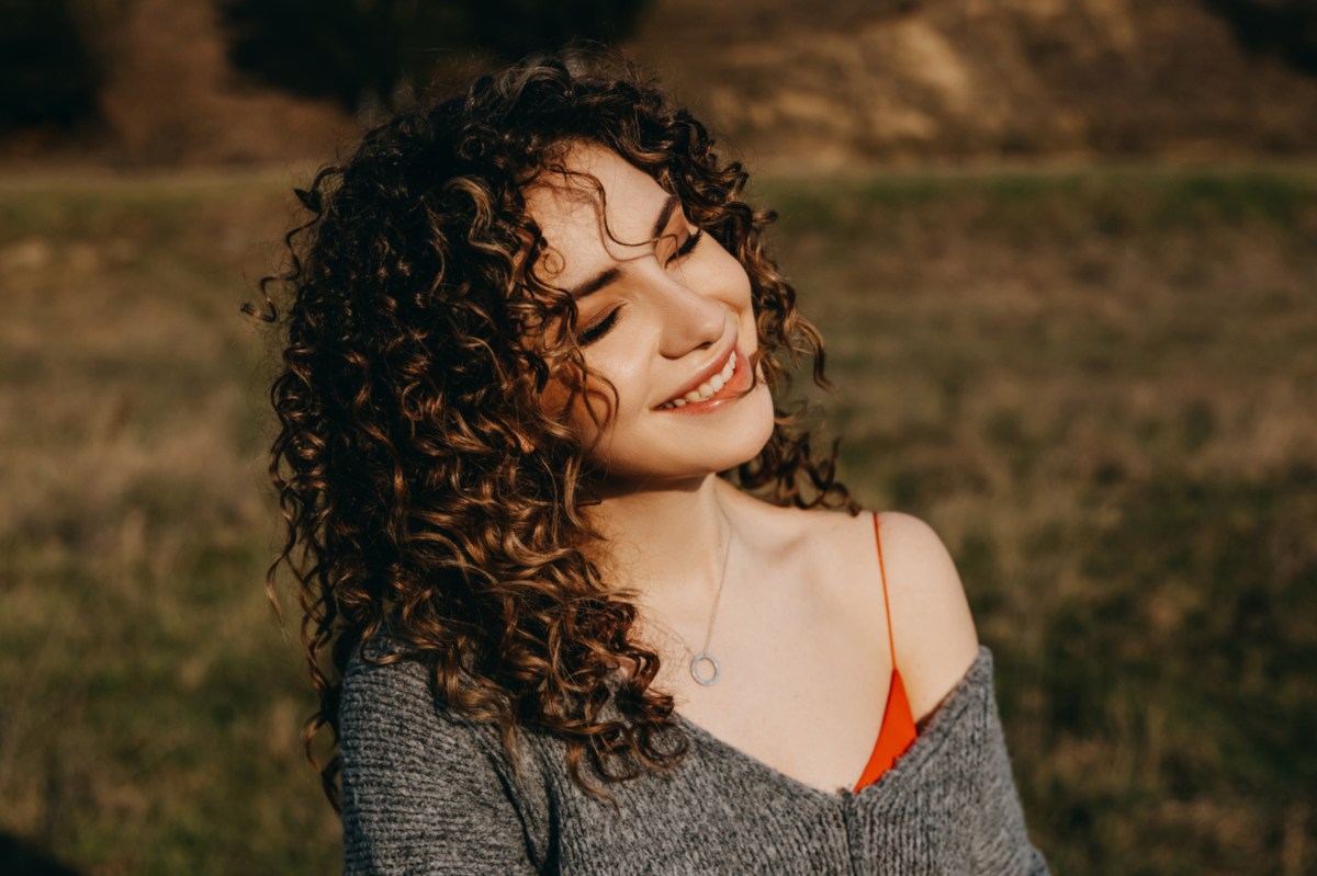 Frau mit lockigem Haar steht in der Sonne auf einer Wiese und lächelt.